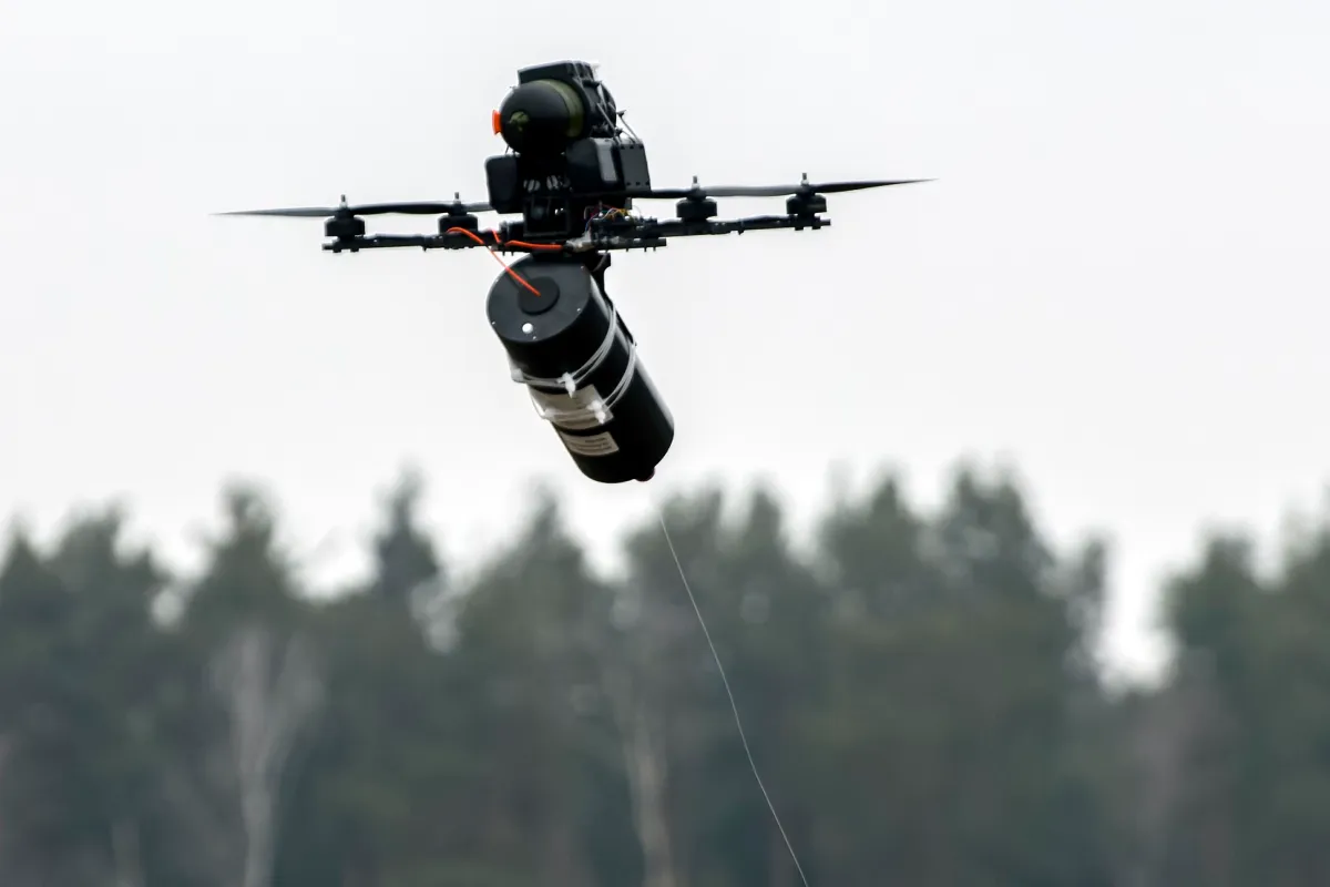 A multirotor drone in flight carrying a munition payload, framed against a treeline.